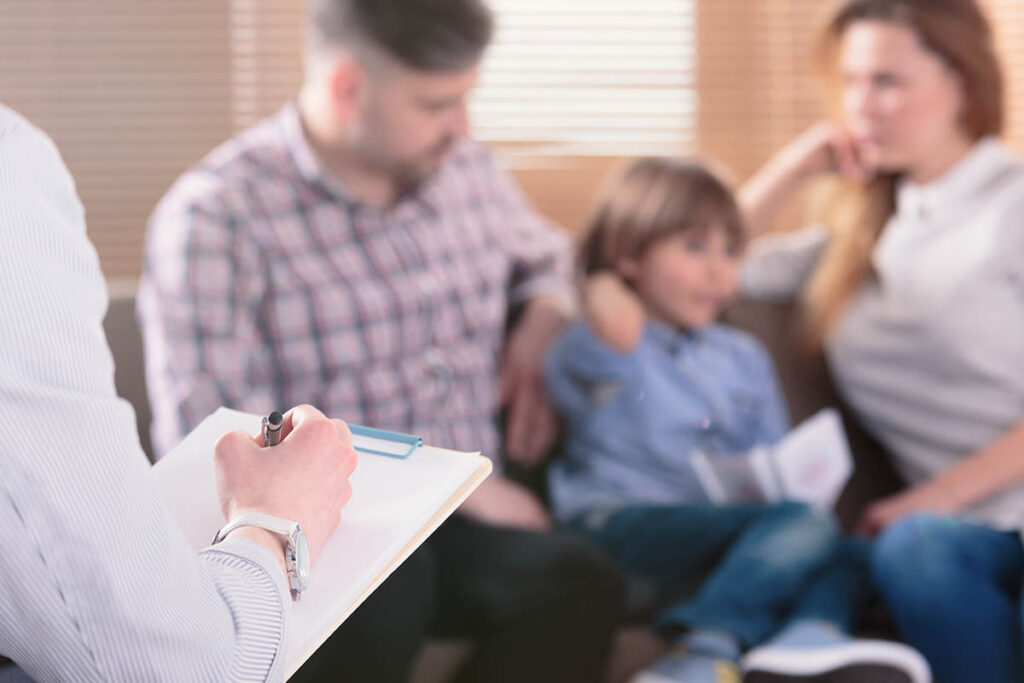 therapist writing on clipboard during a group session where he explains the benefits of family therapy and addiction treatment